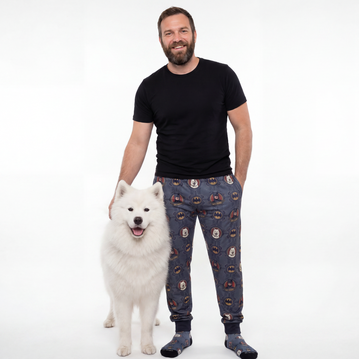 Man wearing a black t-shirt and Dark Knight patterned sweatpants standing next to a white dog on a white background