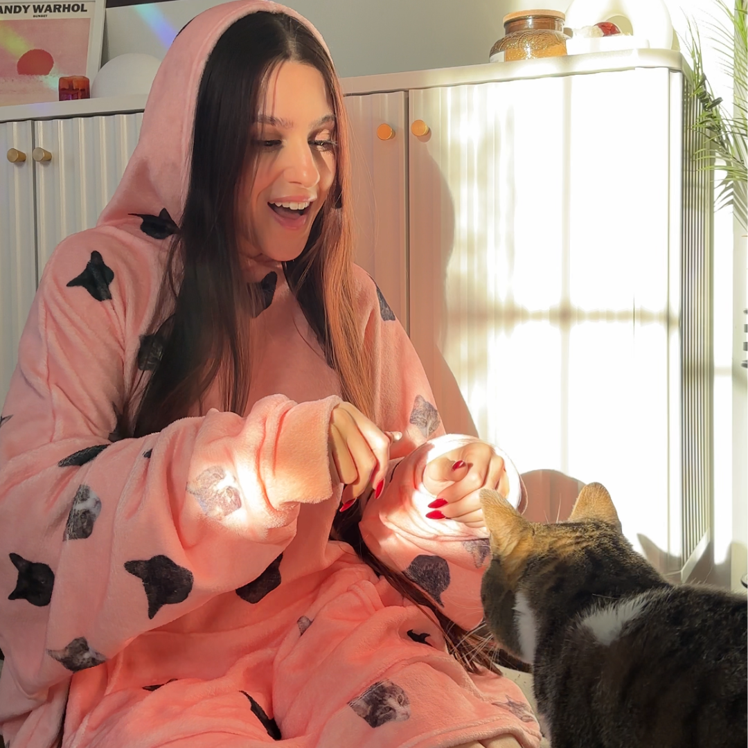 Woman wearing a Pink Dream cat-print hooded robe sitting on the floor and playing with a tabby cat in warm sunlight.