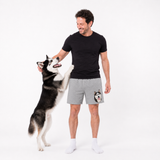 Man wearing a black t-shirt and gray shorts with a dog graphic, standing next to a dog on a white background