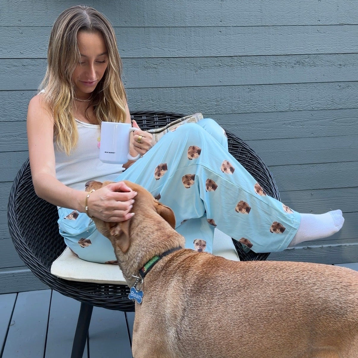Woman wearing sky blue dog-print pajama pants sitting on a patio chair with a mug and petting a tan dog.
