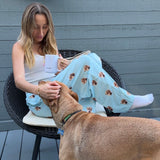 Woman wearing sky blue dog-print pajama pants sitting on a patio chair with a mug and petting a tan dog.
