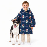 A young boy wearing a navy blue hooded sweatshirt with a festive Christmas dog print stands next to a black, white, and tan dog on a solid white background.