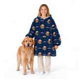 A young girl stands next to a Golden Retriever on a white studio background, wearing a navy blue fleece hooded sweatshirt with an allover Christmas dog print. The print features Santa Claus dogs, snowflakes, and holiday motifs.