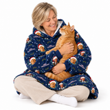 A woman sitting cross-legged on a white studio background, hugging an orange tabby cat while wearing a navy blue hooded pajama set printed with gingerbread cookies, Santa hats, and the words 'Mistletoom Christmas.'.