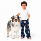 A young boy wearing a white t-shirt and navy blue Christmas-themed pajama pants stands next to a fluffy Australian Shepherd dog on a solid white background.