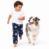 A young boy wearing a white t-shirt and navy blue Christmas pajama pants with a dog print, running barefoot next to an Australian Shepherd on a white studio background.
