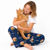 A young girl with blonde hair sits cross-legged on a white studio background, smiling and hugging an orange tabby cat. She is wearing navy blue pajama pants with a festive cat-and-holiday print and a white t-shirt.