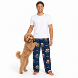 A man wearing a white t-shirt and dark blue Christmas-themed pajama pants stands next to a small golden dog on a white background.