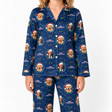 A model wearing a navy blue long-sleeve pajama set with a repeating Christmas-themed print of a golden retriever dog wearing a Santa hat, surrounded by holly leaves and snowflakes. The pajama top has a button-down front, and the matching pants are shown against a plain white background.
