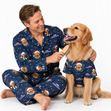A man and a golden retriever dog wearing matching navy blue Christmas-themed pajamas with printed dog portraits and Santa hats, sitting on a white studio background while the man brushes the dog.
