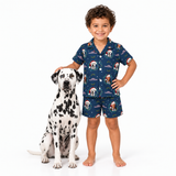 A young boy wearing a navy blue pajama set with a Dalmatian dog print stands next to a real Dalmatian dog on a white studio background.