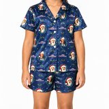 A model wearing a navy blue pajama set with a Christmas-themed print featuring a dog in a Santa hat, the text 'CHRISTMAS KAYAN', and holly motifs. The set includes a short-sleeve button-up shirt and matching shorts against a plain white background.