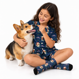 A young girl with long brown hair sits cross-legged on a white studio floor, hugging a tricolor Corgi while wearing a navy blue pajama set with a Santa Claus and Christmas dog print. She is smiling and offering a small treat.