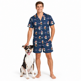 A man wearing a navy blue short-sleeve pajama shirt and matching shorts with a Christmas dog print, standing barefoot next to a black and white dog on a white background.