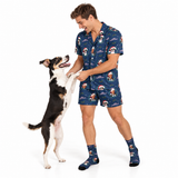 A man wearing a matching navy blue pajama set with a Santa Claus and dog print stands on a white studio background while playfully interacting with a black and white dog. He is also wearing matching socks.