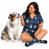 A woman wearing a navy blue Christmas-themed pajama set with a dog print sits on the floor next to an Australian Shepherd dog, offering it a treat against a white studio background.