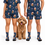 A man and a woman modeling matching navy blue Christmas-themed pajama set with a dog print, paired with matching socks, standing next to a fluffy golden dog on a white background.