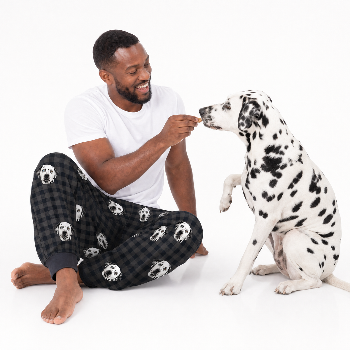 Man in sweatpants with dog pattern feeding a Dalmatian dog on a white background