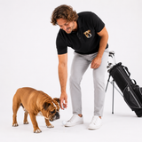 A man in a black polo shirt with a dog logo and light gray trousers bends down to pet a brown English Bulldog on a white studio background, with a black golf bag standing nearby.