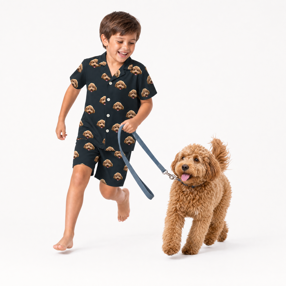 Child wearing a black dog-print pajama short set walking a small tan dog on a leash.