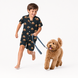 Child wearing a black dog-print pajama short set walking a small tan dog on a leash.