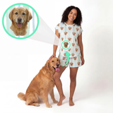 A woman wearing a matching pajama set stands barefoot on a white studio background next to a golden retriever dog. The pajamas are white with light blue vertical stripes and a repeating print of a dog face.
