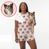 A woman wearing a light pink pajama set with an all-over dog face print holds a small tan and white Chihuahua dog against a plain white studio background. A circular inset at the top left shows a close-up of the dog face that appears in the pajama print.