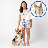 A woman stands barefoot on a white studio background wearing a short-sleeve pajama romper with a corgi print. A corgi dog sits at her left, and circular inset graphics show a close-up of the dog's face and a matching printed detail on the garment.