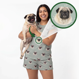 A woman holding a pug dog wears a matching pajama set with a custom printed design of the dog's face. The set consists of a light sage green short-sleeve top and shorts with a repeating pattern of the pug's face.