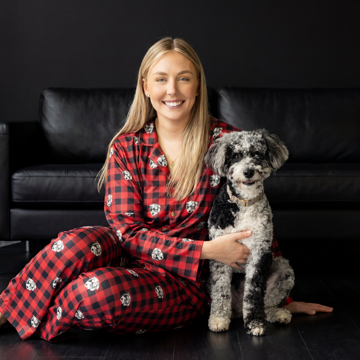 Woman in red and black checkered pajamas holding a small dog on a dark couch.