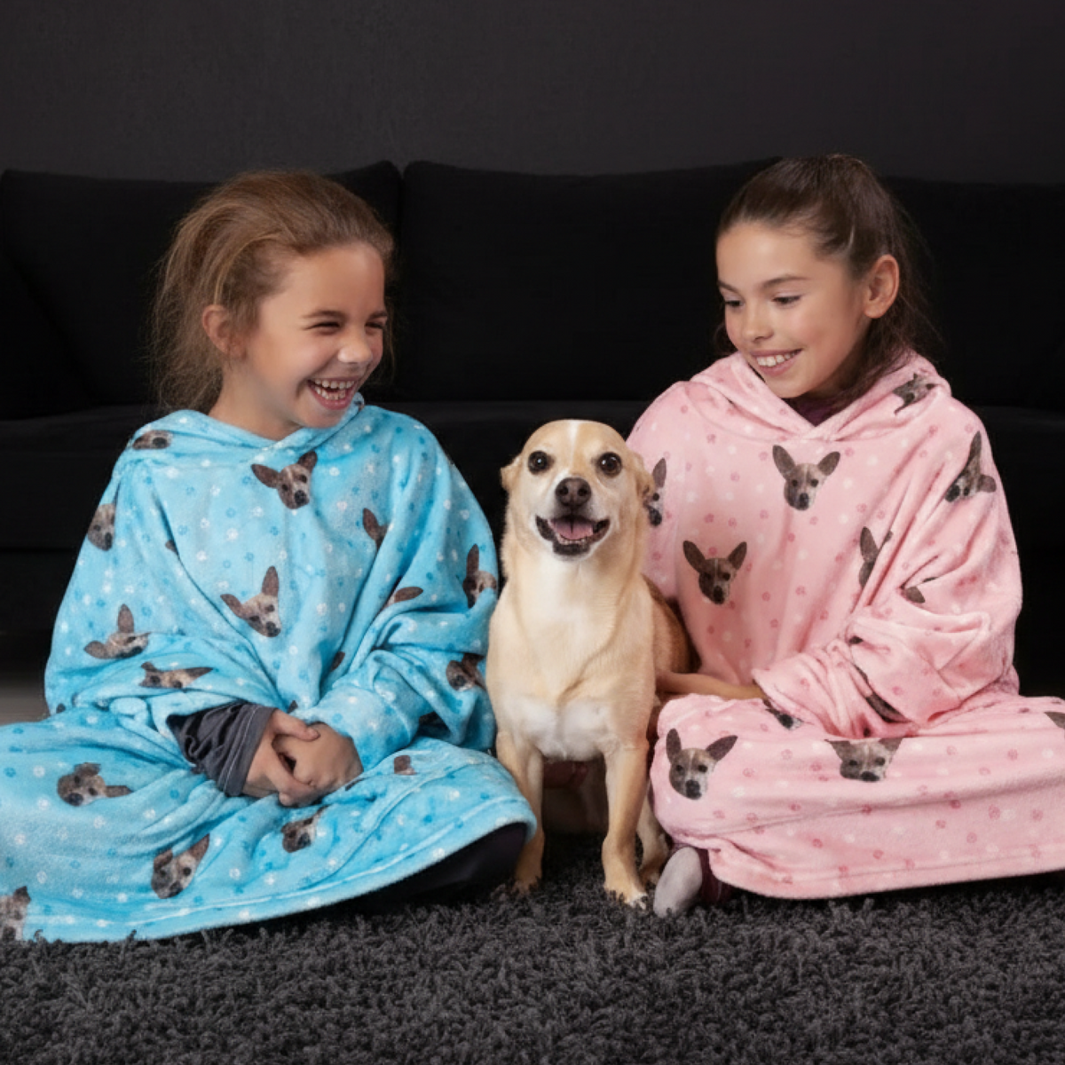 Two children in animal-themed hooded blankets sitting with a dog on a dark floor.