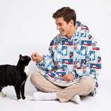 A man wearing a blue and white cat-print hooded blanket jacket sits cross-legged on a white studio floor, smiling at a black and white cat.
