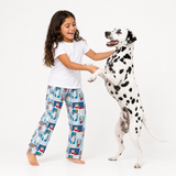 A young girl with long dark hair smiles as she pets a Dalmatian dog standing on its hind legs. The girl is wearing a white t-shirt and light blue pajama pants with a cartoon print, while the dog wears a white coat with black spots.