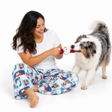 A woman sitting on the floor plays with a dog tug-of-war toy with her dog. She is wearing a white t-shirt and blue patterned pajama pants, and the dog is a blue merle with white and tan markings.