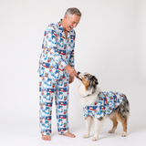 A man and a dog wearing matching blue and white dog-themed pajamas, standing on a light gray studio background. The man is barefoot, and the dog is wearing a matching dog pajama top.