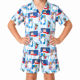 A child wearing a matching short-sleeve button-up pajama shirt and shorts set with a winter dog-themed print. The pajamas feature a light blue background with a collage of cartoon dogs in snow hats and scarves, along with snowman motifs, on a plain white studio background.