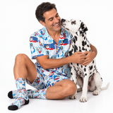 A man sitting on a white studio floor wearing a matching Dalmatian-themed short-sleeve pajama shirt, shorts, and socks, hugging a Dalmatian dog.