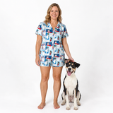 A woman stands barefoot on a white studio background wearing a short-sleeve pajama set with a blue, white, and red patchwork-style print. A black-and-white dog sits beside her, and the product photo is clearly intended to show the pajamas being worn.