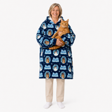 A woman stands against a light gray studio background wearing a navy blue hooded robe with a repeating 'Fuzzy Pups' dog print, while holding an orange tabby cat. The robe has a soft plush texture and the print includes white, light blue, and tan details.