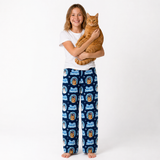 A young girl with blonde hair stands barefoot against a white studio background, holding an orange tabby cat. She is wearing a white t-shirt and navy blue pajama pants with a repeating cartoon dog pattern and the text 'Just Dog'.