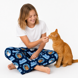 A young girl sitting on a white floor, smiling as she feeds an orange tabby cat. She is wearing a white t-shirt and navy blue pajama pants with a 'Frozen' and 'Snow White' cat-themed pattern.