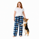 A woman stands barefoot against a white studio background wearing navy blue pajama pants with a repeating 'Just Do It' dog graphic print, paired with a plain white t-shirt. A small black, white, and tan dog stands beside her, touching her arm.