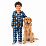 A young boy wearing a blue long-sleeve pajama set with a repeating cartoon dog pattern stands barefoot next to a golden retriever dog on a plain white studio background.