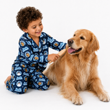 A young boy with curly brown hair wearing blue and white patterned pajamas kneels on a white studio floor, brushing a golden retriever dog with a wooden-handled brush.