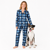 A young girl wearing a navy blue long-sleeve pajama set with a repeating cartoon dog pattern stands barefoot next to a black, white, and tan dog on a plain white studio background.