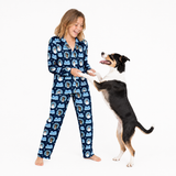 A young girl wearing a navy blue long-sleeve pajama set with a repeating cartoon dog pattern, standing barefoot on a white background and playfully reaching toward a black, white, and tan dog.