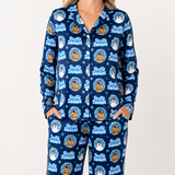 A woman wearing a matching long-sleeve button-front pajama top and pants set in navy blue with an allover 'Frosty Snowshoe' cartoon print. The print features orange dogs in snow boots inside blue circular frames, along with snowmen and white snowflakes.