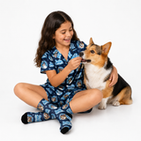 A young girl with long dark hair sits cross-legged on a white studio background, smiling as she feeds a corgi dog. She is wearing a navy blue pajama set with a repeating cartoon dog face pattern and matching socks.