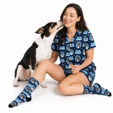 A woman and a black-and-white dog wearing matching navy blue pajama set with a cartoon dog print. The set includes a short-sleeve button-up top and shorts, along with matching socks.