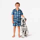 A young boy wearing a blue and white printed short-sleeve pajama set stands barefoot next to a Dalmatian dog on a clean white studio background. The pajama set features a repeating pattern of cartoon characters and the word 'Pajama'.